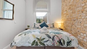 A bedroom with a large bed, a brick wall, a window with blinds, and a mirror on the wall. - Apartment 1 - The Coachhouse (Sydney)