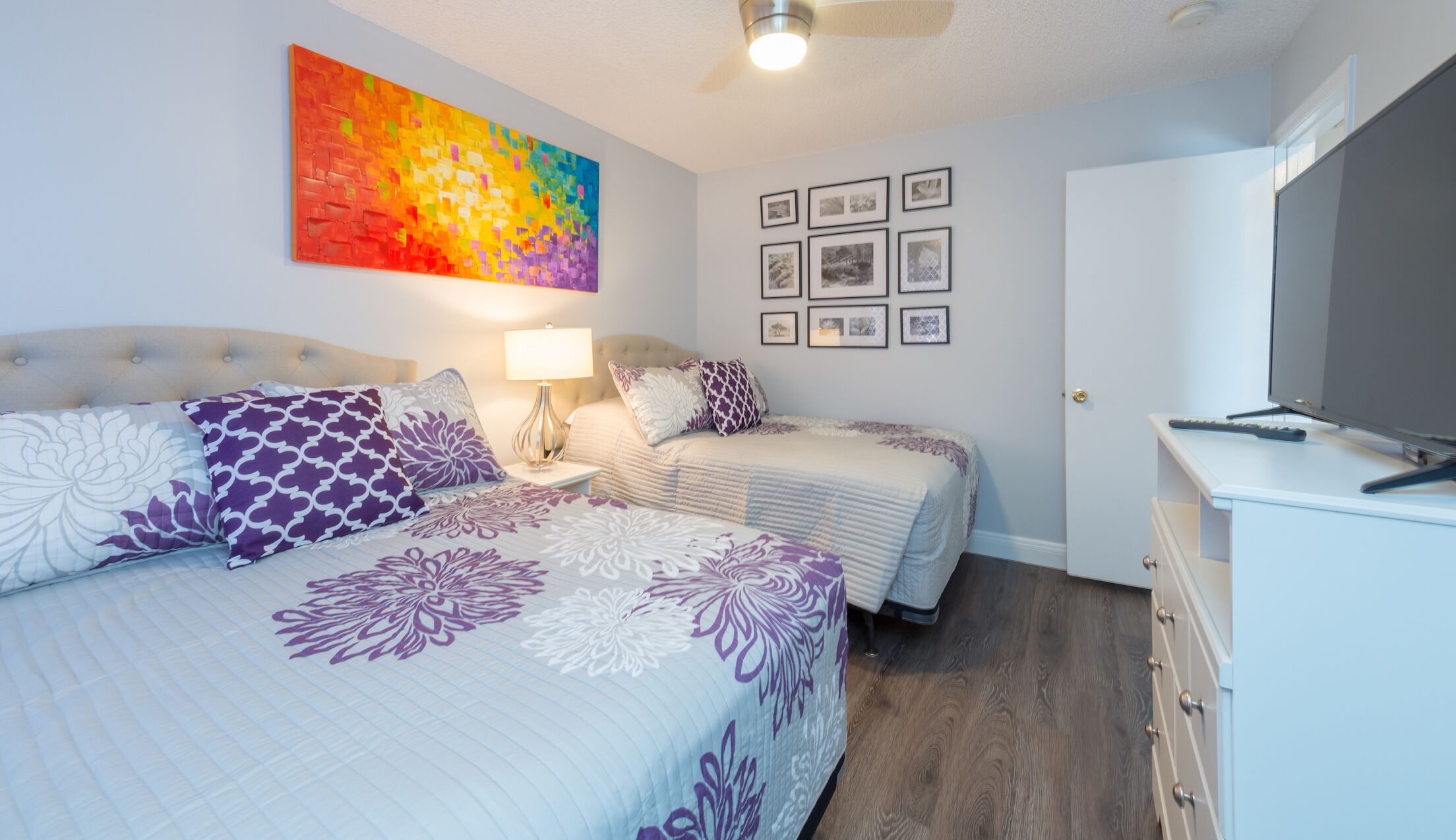 A bedroom with two beds, a TV, a desk, and a colorful painting on the wall.