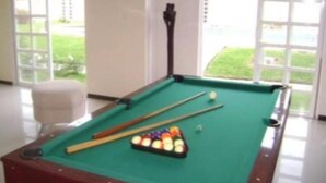 A pool table with pool cues and balls, a white chair, and large windows with a view of greenery. - Casa Familiar Acapulco Punta Diamante, Fracc. Puente del Mar, cerca de la playa. (Aeropuerto)