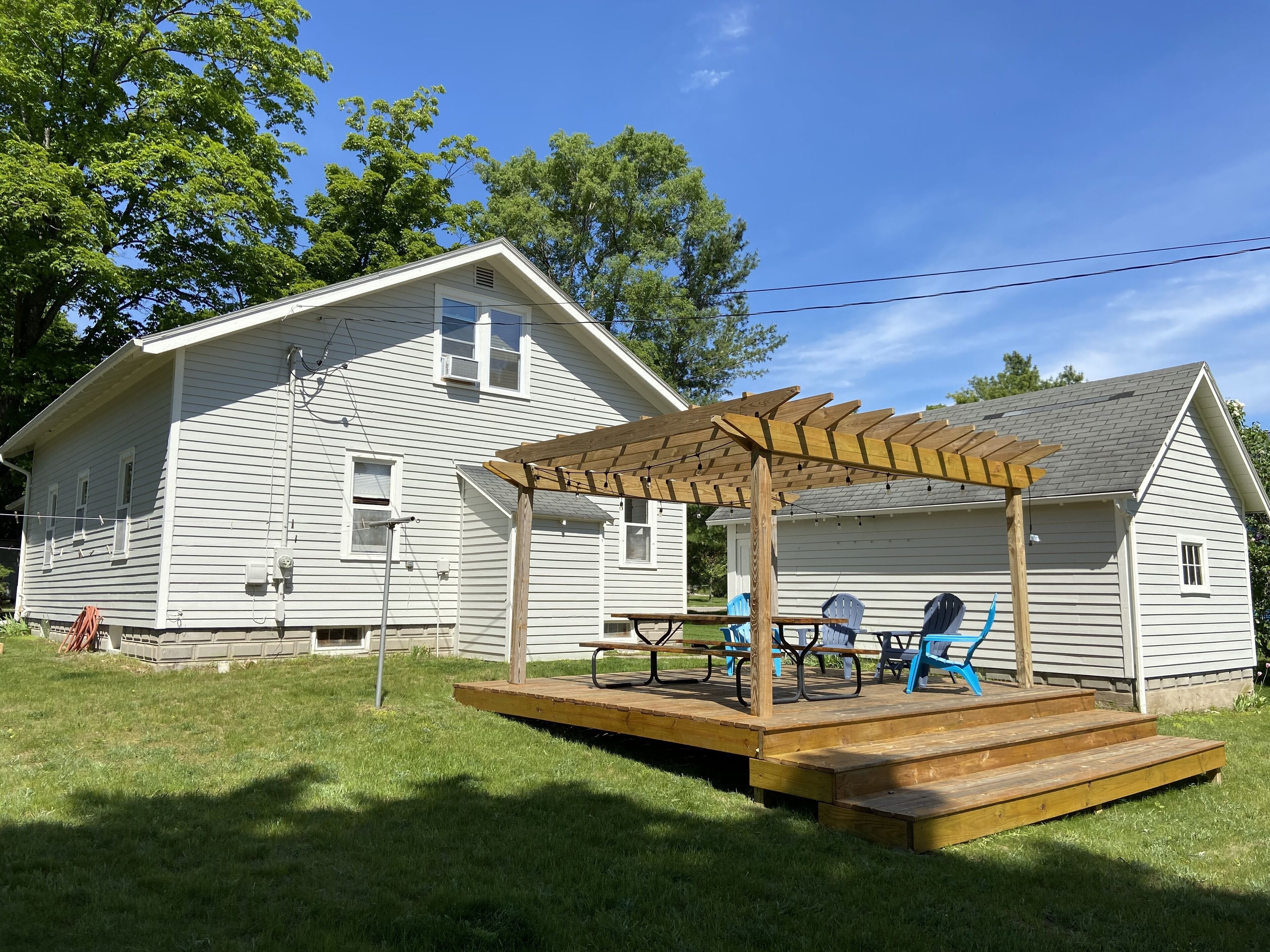 En träveranda med en pergola, ett picknickbord och blå stolar framför ett vitt hus med ett sluttande tak.