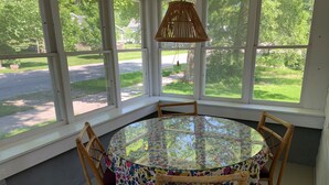 A round glass table with a floral tablecloth, surrounded by four wicker chairs, set in a room with large windows offering a view of a green landscape and a road. - Home located 2 blocks to the beach and Suttons Bay Village (Suttons Bay)