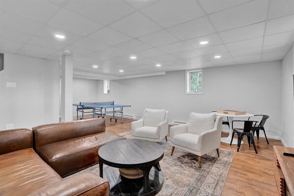 A modern living room with a brown leather sofa, white armchairs, a round coffee table, and a ping pong table in the background.