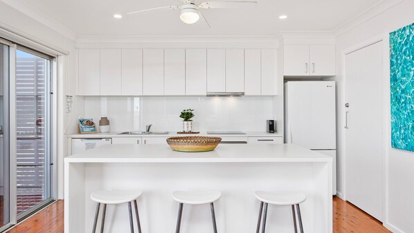 A modern kitchen with white cabinetry, a central island with stools, a ceiling fan, and a large window with blinds. - Spirit of Gerroa - Moments from Seven Mile Beach (Gerroa)