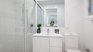 A modern bathroom with a glass shower enclosure, a white sink with two black soap dispensers, and a small potted plant. - Spirit of Gerroa  - Moments from Seven Mile Beach (Gerroa)