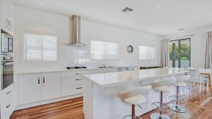 Une cuisine moderne avec des meubles blancs, un îlot central doté de tabourets et de grandes fenêtres équipées de stores.