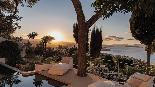 Uma área de estar tranquila ao ar livre com piscina, cercada por árvores e com vista panorâmica para o mar e montanhas ao longe.