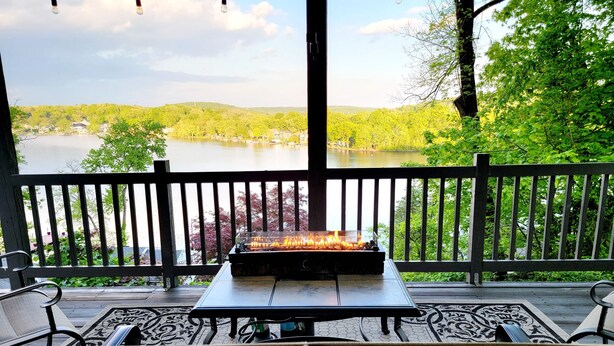 Un espace extérieur aménagé, abrité et doté d’un foyer, offrant une vue sur un lac et des collines boisées.