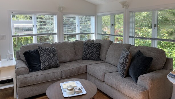 A modern living room with a grey sectional sofa, a round coffee table with books and a candle, and large windows with blinds.