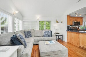 A modern living room with a grey sectional sofa, a small side table, and a kitchen area with wooden cabinets and a microwave.