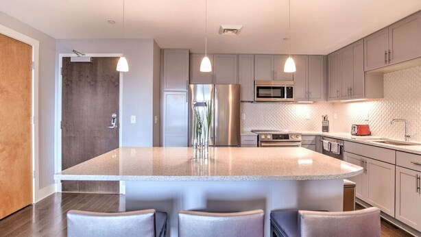 Une cuisine moderne dotée d’un grand îlot central, d’appareils électroménagers en acier inoxydable et de luminaires suspendus.