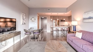 Un salon moderne avec un canapé, une table basse et un téléviseur. Au fond, on aperçoit une cuisine avec un coin repas.