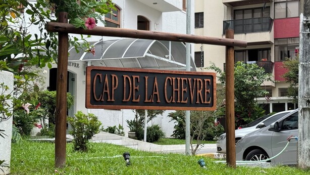 Een bord met 'CAP DE LA CHEVRE' in een rustieke houten lijst, omringd door groen en een modern gebouw op de achtergrond.