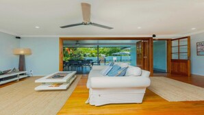 A living room with a white sofa, a coffee table, and a ceiling fan. There is a door leading to an outdoor area with a view of greenery. - TROPICAL ESSENCE @ PALM COVE (PALM COVE)
