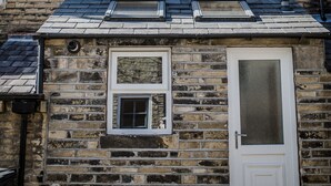 Un bâtiment en pierre avec un toit plat, deux fenêtres avec volets, une porte dotée d’un panneau de verre et une petite lucarne.