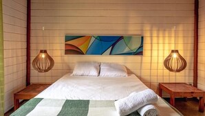 Un dormitorio con una cama, dos mesitas de noche, un cuadro abstracto en la pared y dos lámparas colgantes.
