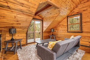 A cozy cabin with a loft bedroom, wooden walls, a sofa, two tables, and a view of trees through large windows.