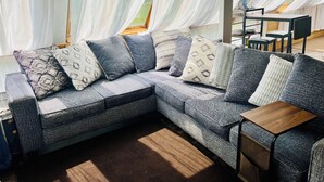 Un salon moderne avec un canapé d’angle gris, des coussins à motifs, une petite table d’appoint en bois et des rideaux blancs transparents.