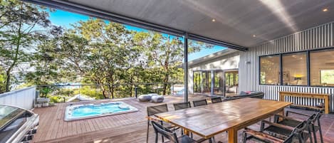 An outdoor deck with a hot tub, wooden table, and chairs, overlooking a forested area.