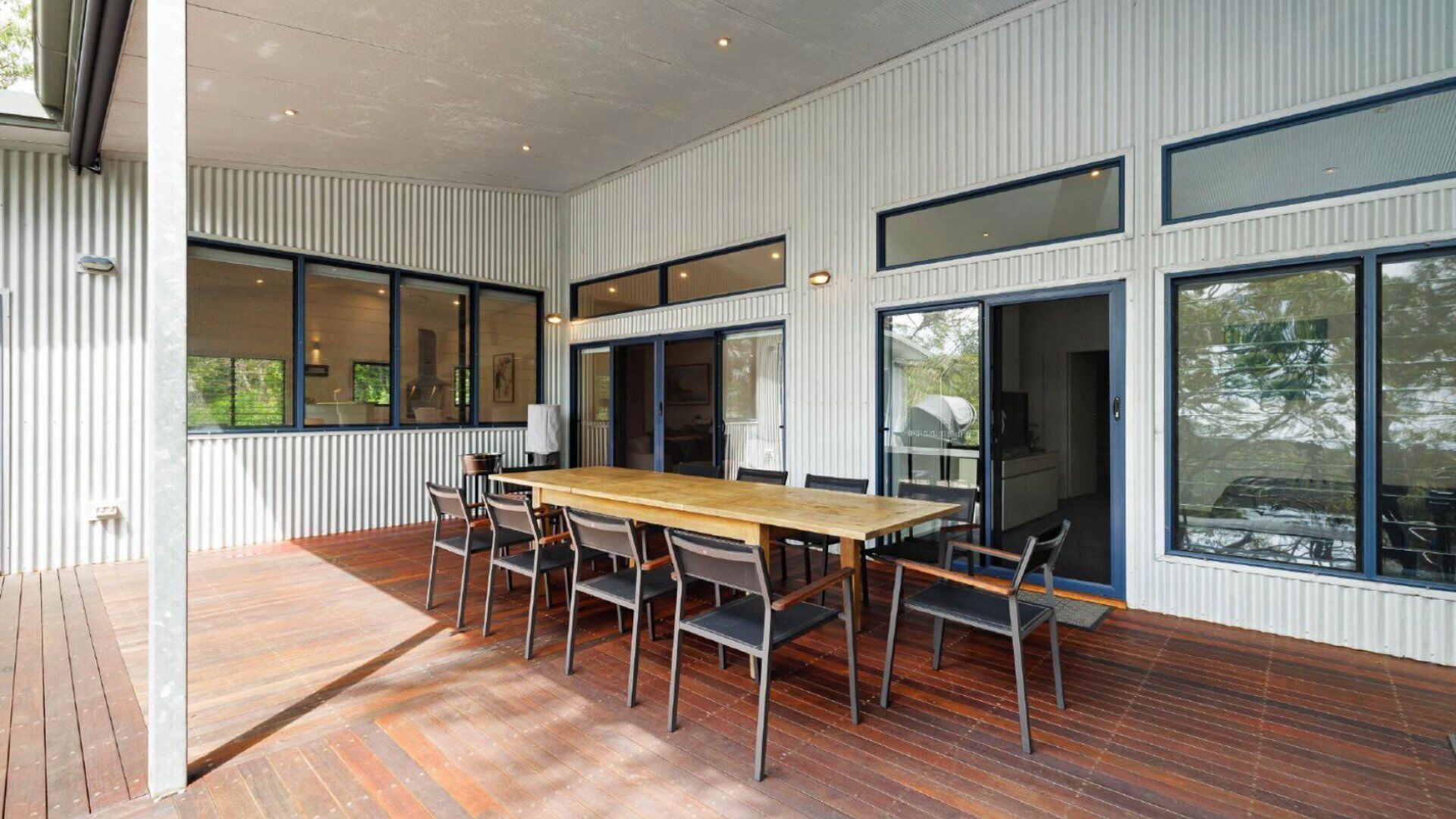 A modern dining area with a wooden table and chairs, large windows, and a view of greenery outside.