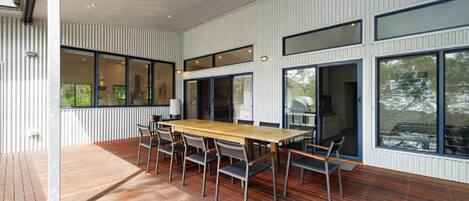 A modern dining area with a wooden table and chairs, large windows, and a view of greenery outside.