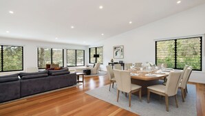 A spacious living area with a sofa, dining table set for six, and large windows offering a view of greenery.