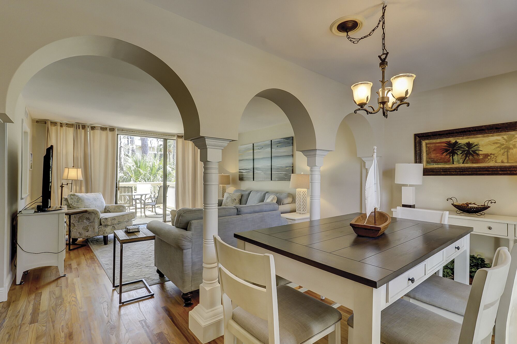 A spacious living area with a dining table, white chairs, and a sofa. There are arched doorways and a chandelier.