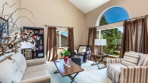 Un salon spacieux comprenant un canapé, un fauteuil et une table basse. On y trouve une bibliothèque avec des objets décoratifs et une fenêtre agrémentée de rideaux.