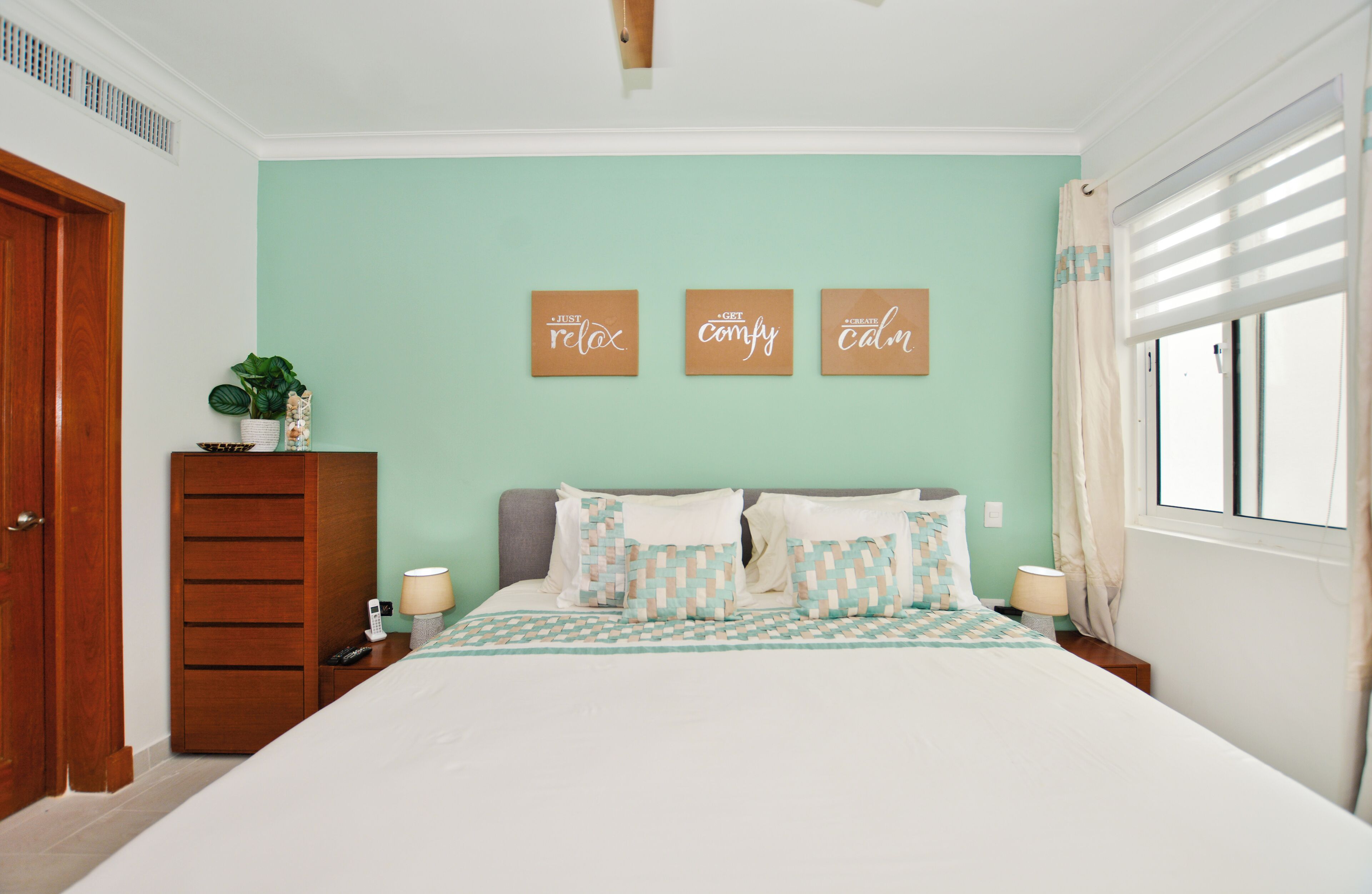Quarto com cama, mesas de cabeceira, penteadeira e um painel com três frases motivacionais.