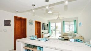 A modern kitchen with white countertops, a central island with blue plates, and a dining area with a sofa and a TV. - Playa Turquesa - B-302 -Premier Beachfront Ocean View 400mbps Wifi &Free Netflix (Punta Cana)