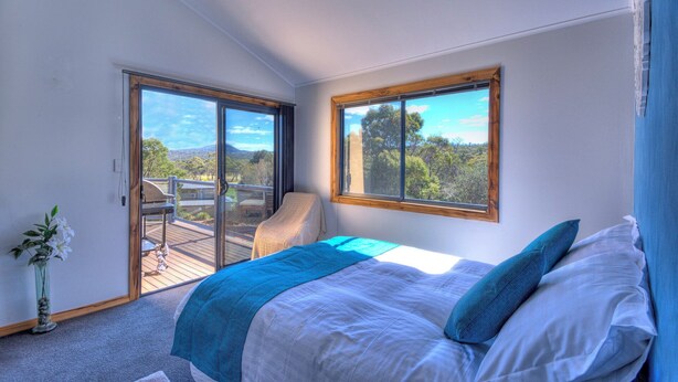 A bedroom with a bed, a chair, a view through a glass door, and a window. - Mayakitana - Freycinet Holiday Houses (Coles Bay)