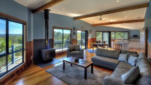 A spacious living room with a large sofa, a coffee table, and a fireplace. There is a sliding glass door leading to a balcony with a view of the outdoors. - Mayakitana - Freycinet Holiday Houses (Coles Bay)