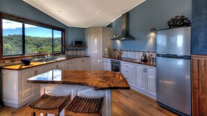 A modern kitchen with a large island, wooden bench seating, and a view of the forest through the window. - Mayakitana - Freycinet Holiday Houses (Coles Bay)
