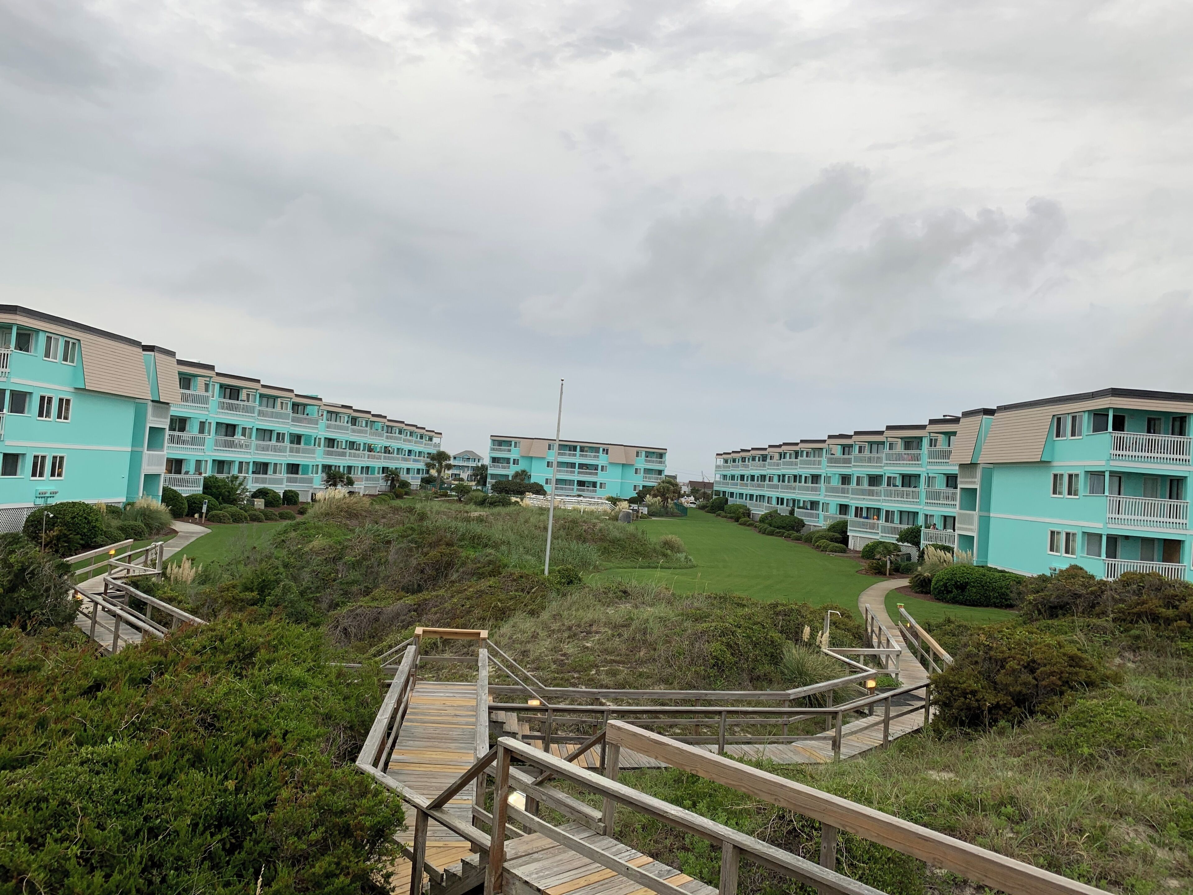 A coastal residential area with multi-story buildings, wooden walkways, and a grassy landscape.