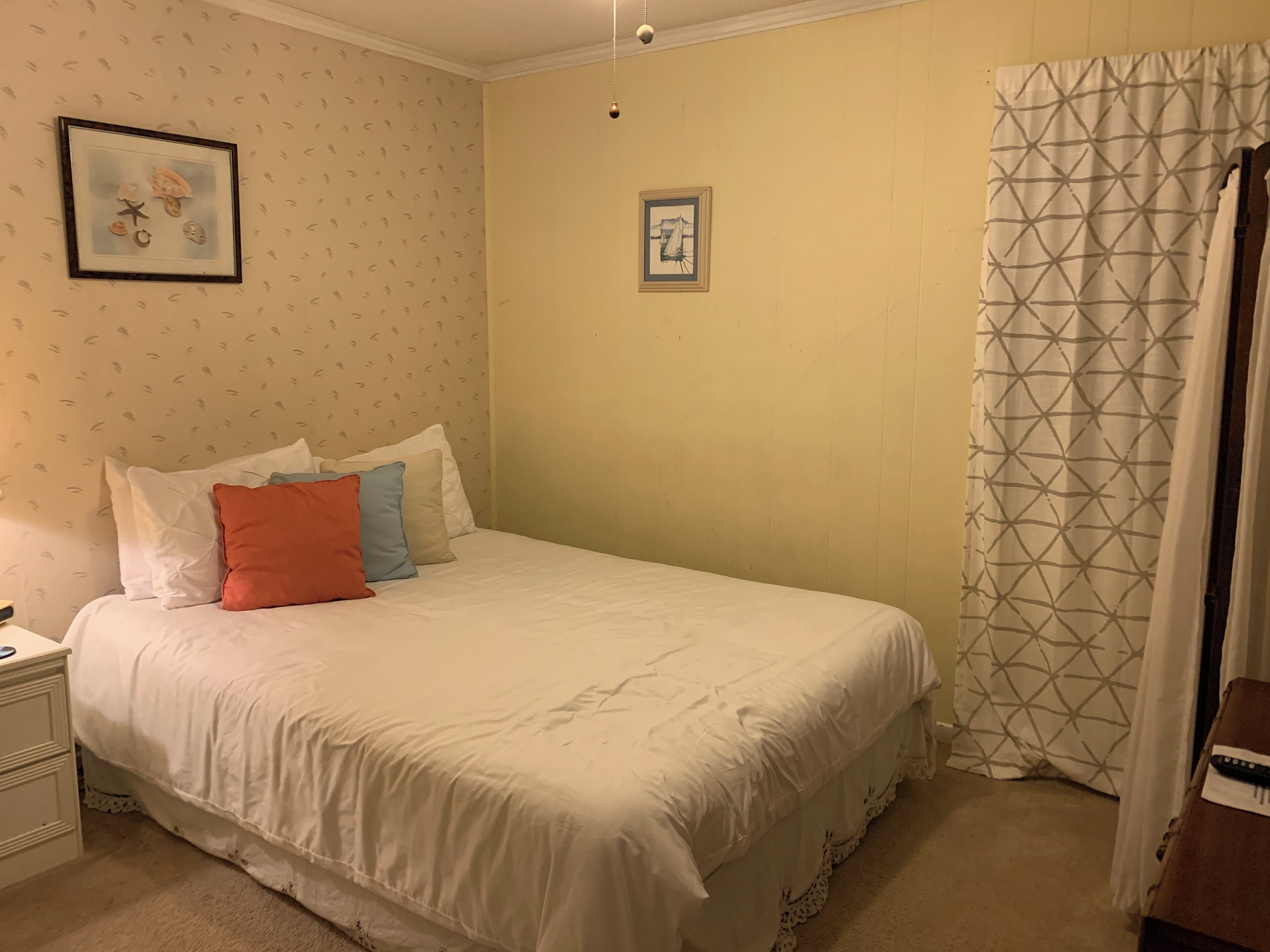 Un dormitorio con cama, almohadas, mesita de noche, un cuadro enmarcado, un reloj de pared y una cortina.