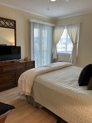 A bedroom with a bed, a television on a wooden dresser, and a window with curtains. - Ocean-side condominium with pool. Fully-equipped for relaxed vacationing. (Atlantic Beach)