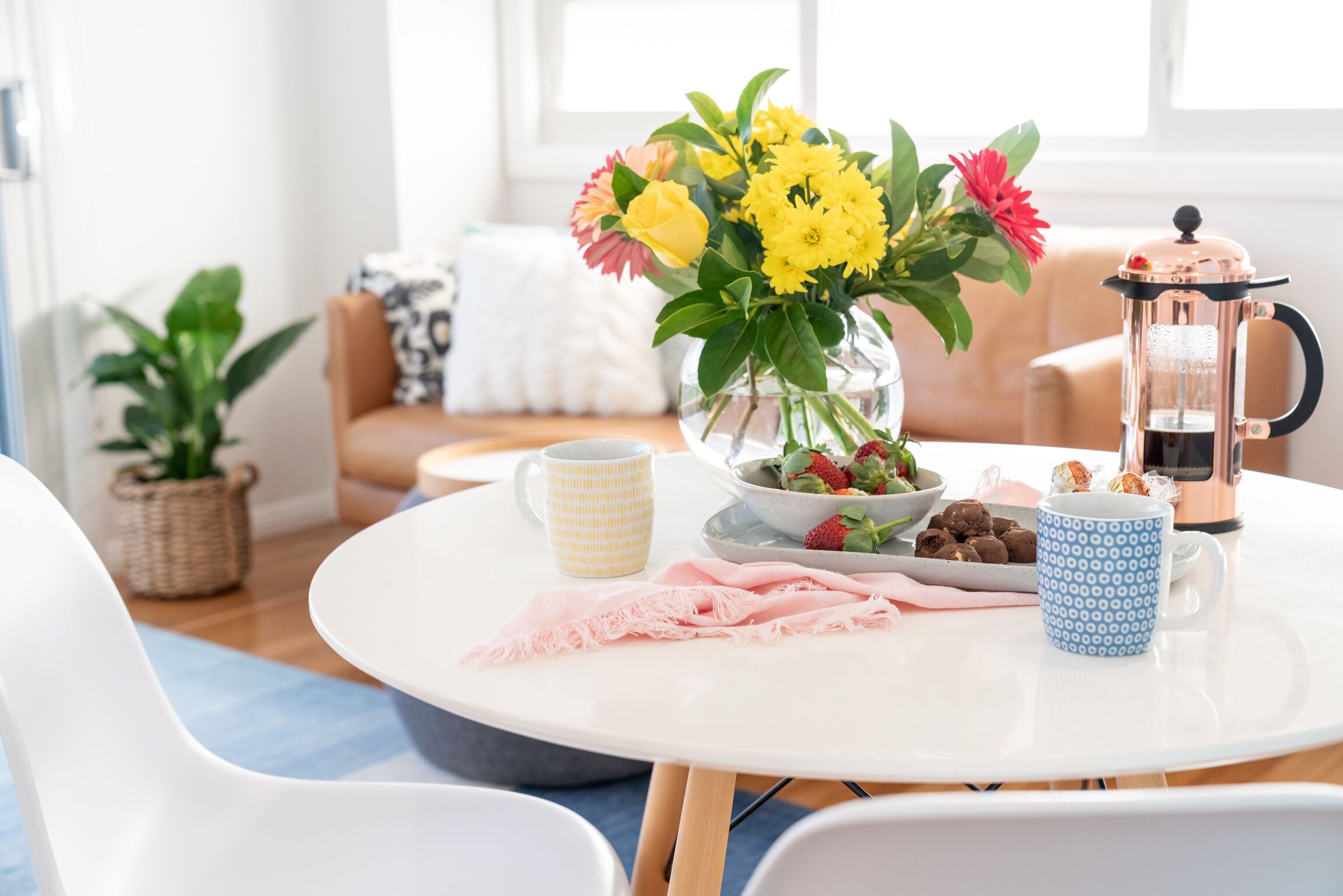 Een ronde witte tafel met een vaas met gele bloemen, een koffiepot en een schaal met aardbeien en chocolade.