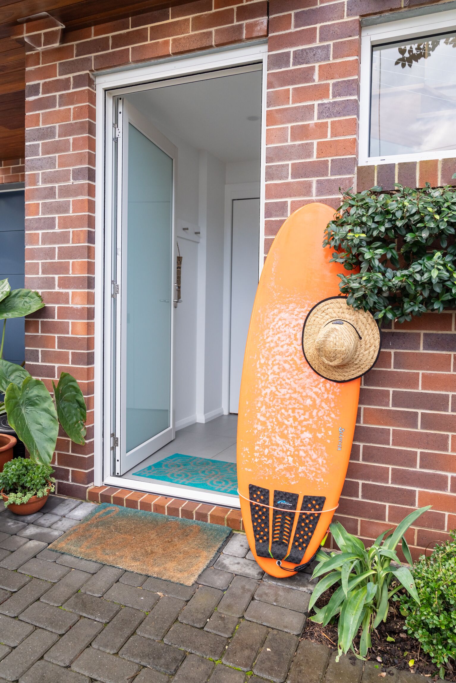 Un edificio de ladrillo con un cerramiento de vidrio correderas que da a una habitación. Una tabla de surf naranja está apoyada contra la pared, y un sombrero de paja encima.