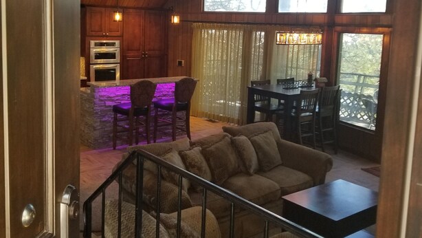 A wooden interior with a staircase leading to a dining area, a kitchen with built-in appliances, and large windows providing natural light. - Surrounded by Pines, Family / Pet Friendly minutes from Snow Valley !!! (Running Springs)