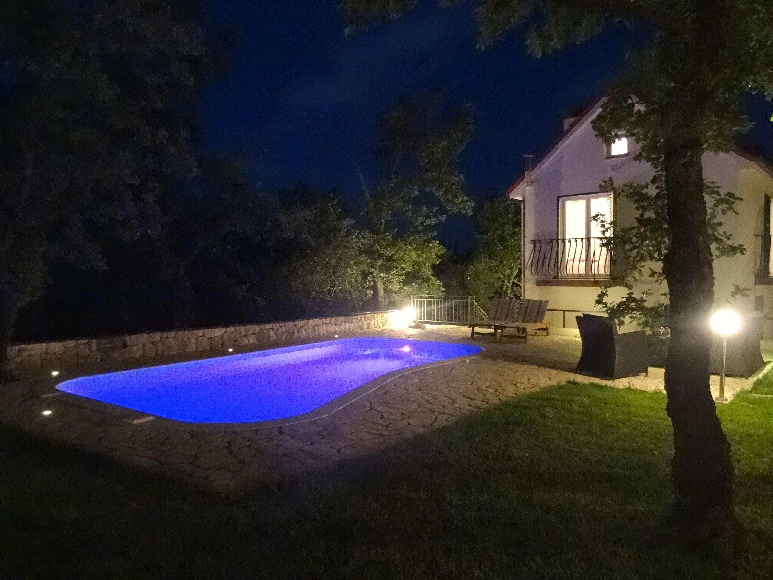 Une piscine bordée de pierres, un mur en pierre et une zone herbeuse avec une maison en arrière-plan.