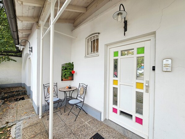 Un bâtiment blanc avec une porte ornée d’une fenêtre aux couleurs verte et jaune, un pot de fleurs et une petite table avec des chaises.