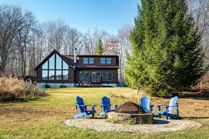 Una casa de dos pisos con un amplio porche con ventanas, un brasero y sillas Adirondack azules.