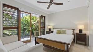 A bedroom with a bed, two bedside tables, a chair, a ceiling fan, and a sliding glass door leading to a balcony with plants. - LARASATI  @ PALM COVE (PALM COVE)