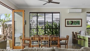 A dining area with a wooden table and chairs, a ceiling fan, and a view through a window with blinds. - LARASATI  @ PALM COVE (PALM COVE)