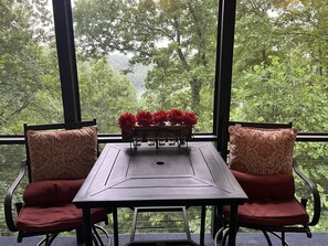 Un tavolo con un vaso di fiori rossi, due sedie decorate e vista su alberi attraverso ampie finestre.
