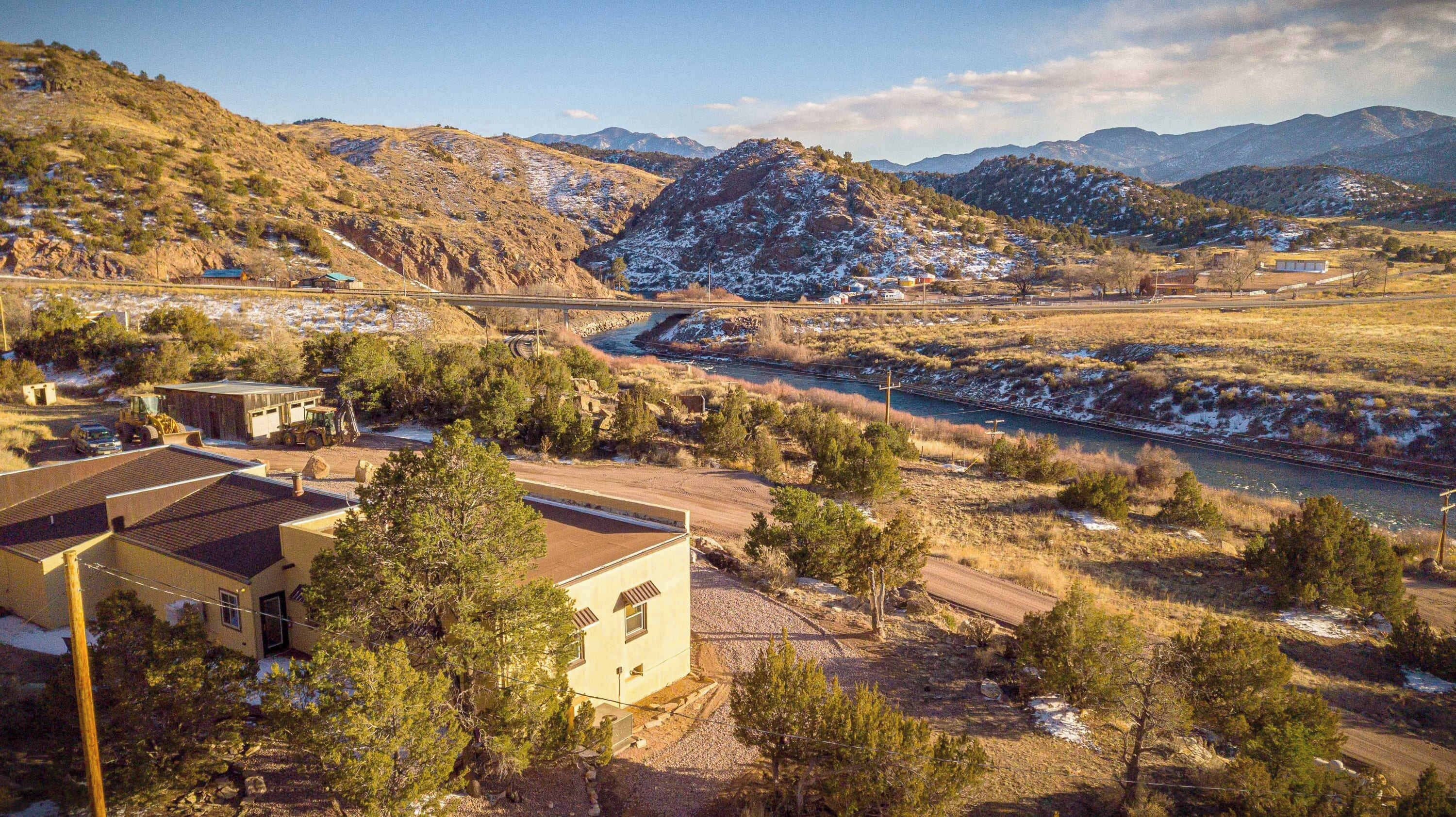 Paisaje rural con un río, montañas y un puente. Se observan edificios y árboles en primer plano.