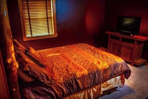 Une chambre à coucher avec un lit, un téléviseur posé sur un meuble en bois et une fenêtre avec des stores.