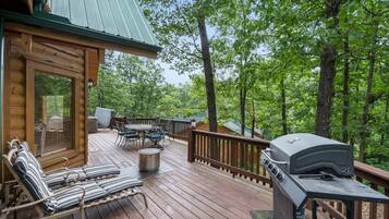 Una terraza de madera con mobiliario exterior, una parrilla y vistas al bosque circundante.