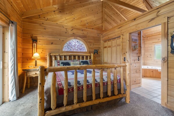 A wooden cabin room with a bed, a desk, a lamp, and a window with a stained glass design. - Refreshed Three Bear Log Cabin, Vaulted Ceilings, Private Hot Tub, Car Charger, Close to Big Cedar (Ridgedale)
