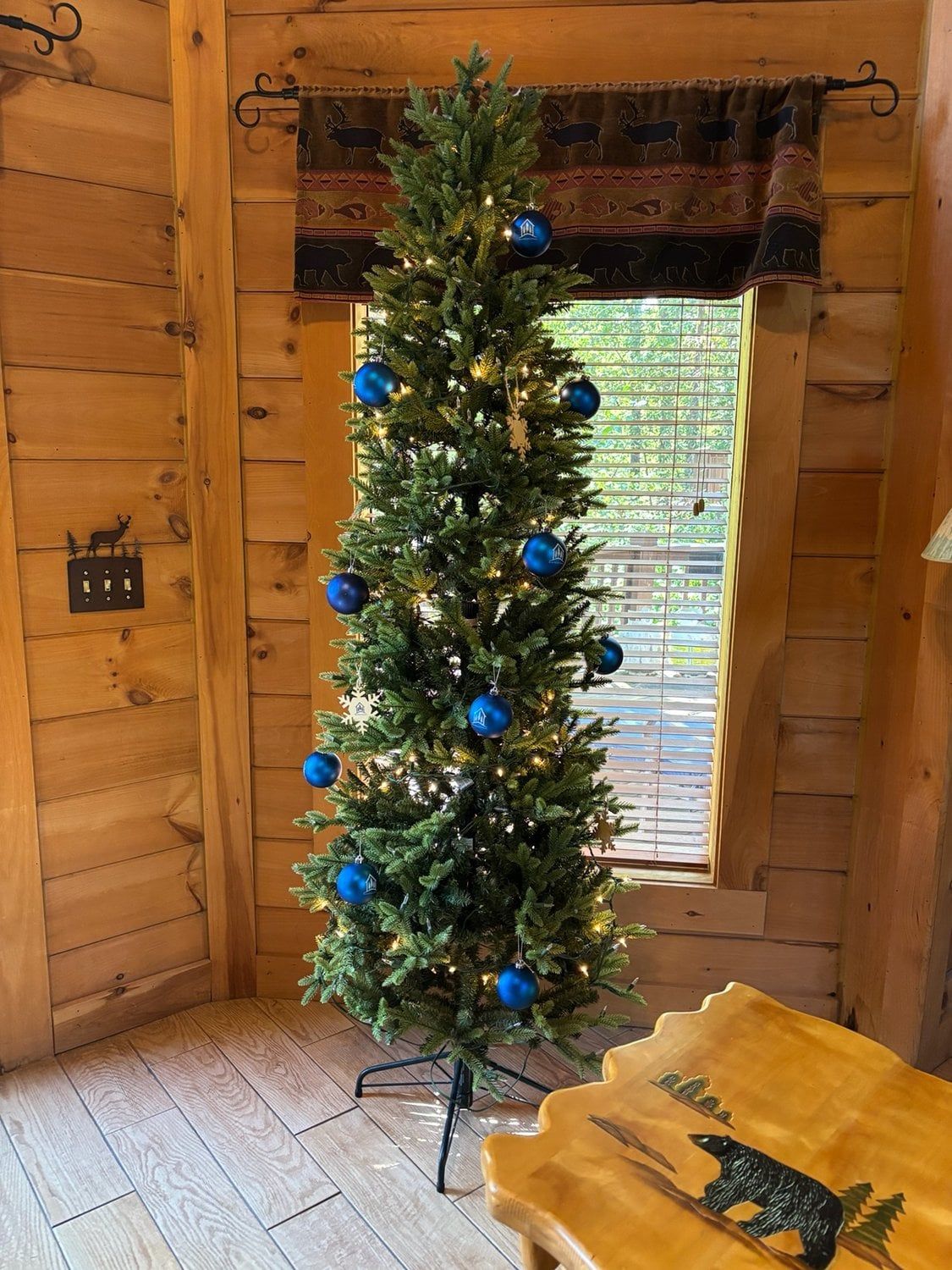 Un árbol de Navidad con adornos azules en una habitación de cabaña de madera con ventana y una mesa con diseño de oso.