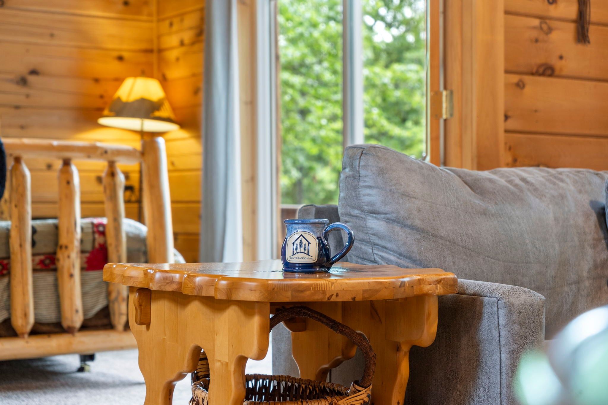 A wooden table with a mug in front of a grey sofa in a room with wooden walls and a window.